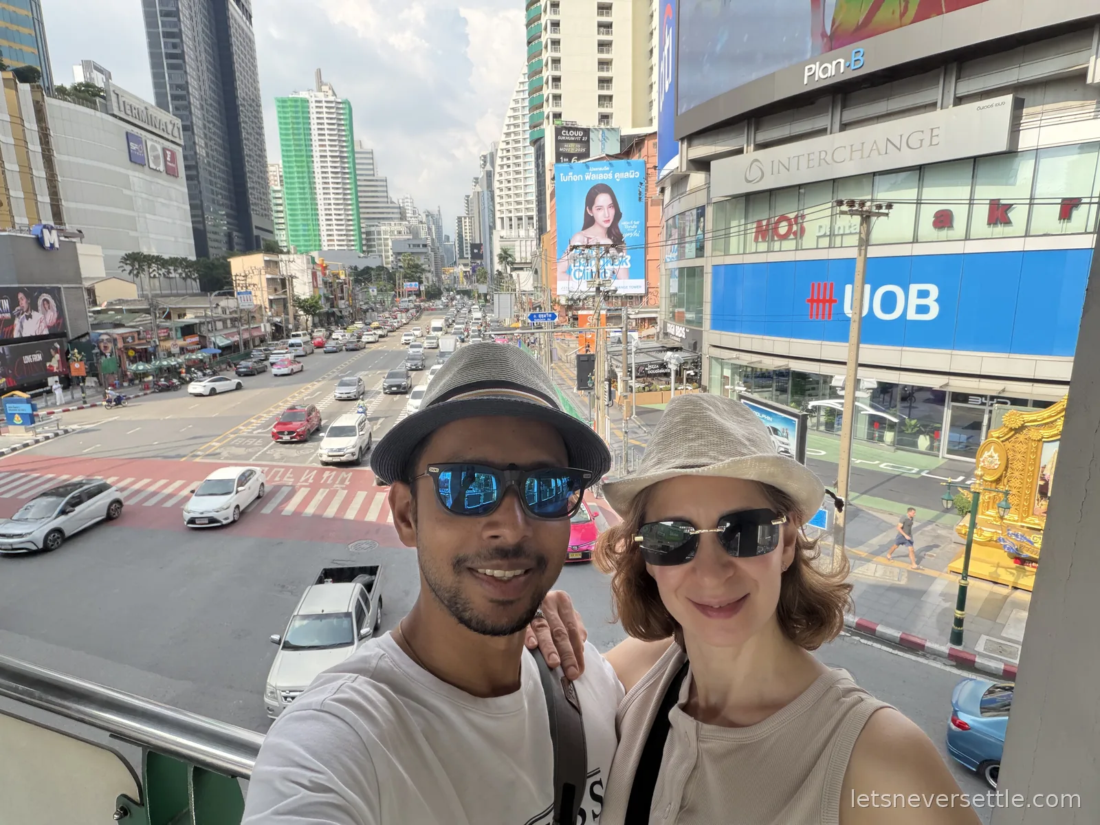 Selfie of Dileep and Inga in sun hats on a skywalk above the Asok intersection, Sukhumvit Road traffic and Terminal 21 behind