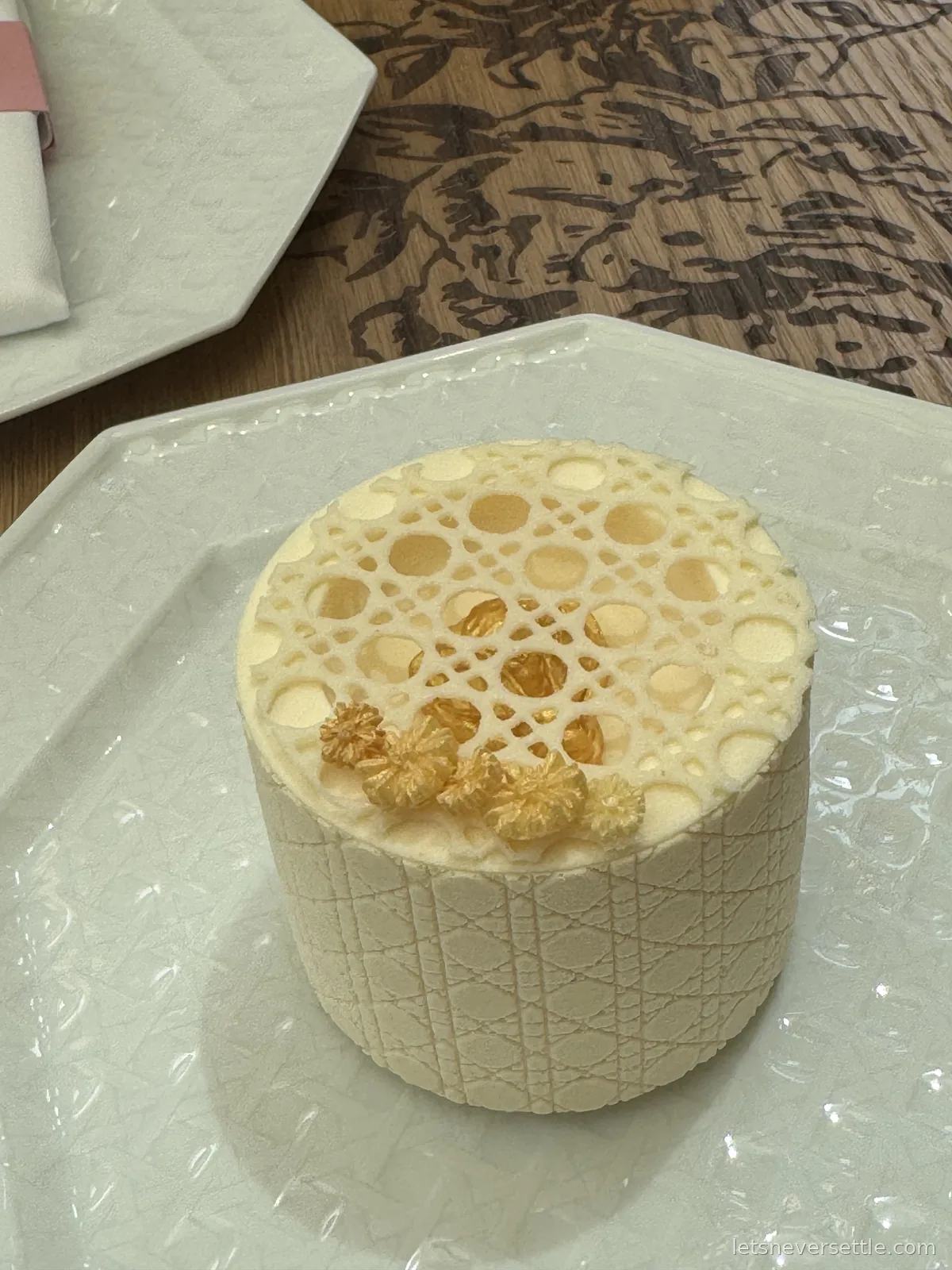 A small cylindrical cream-coloured cake with a woven cannage pattern down the sides, a lattice glaze on top and gold sugar flowers