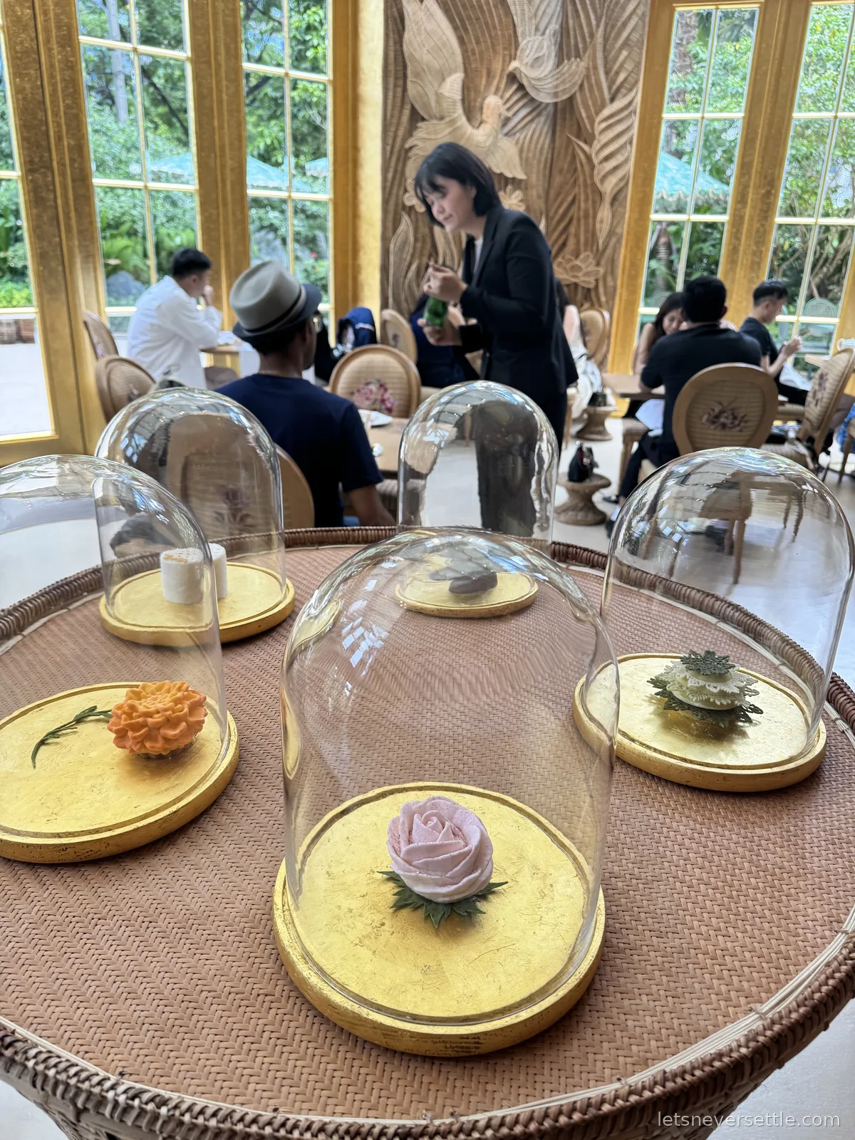 Glass cloches on gold-leaf bases on a rattan table, covering a pink sugar rose, an orange marigold and a green pastry, with tall gilded windows behind