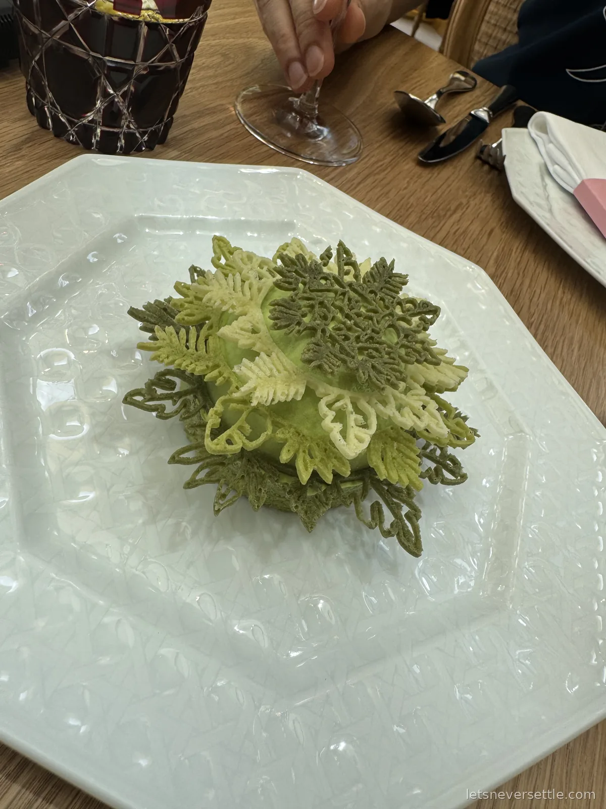 A green matcha dessert sculpted into layered leaves and fronds, sitting alone in the middle of a large white octagonal plate
