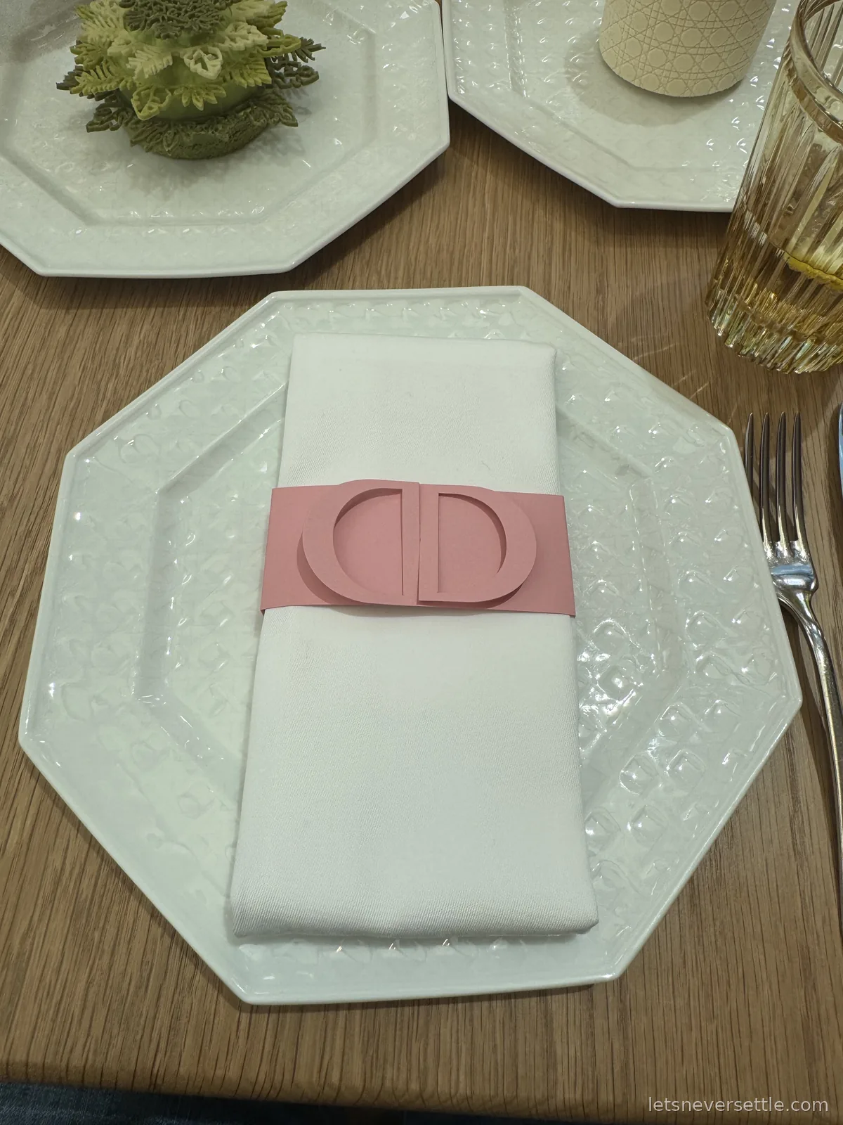 A white embossed octagonal plate with a folded napkin held by a pink paper band printed with the interlocking CD monogram, beside a fork and a gold-rimmed glass