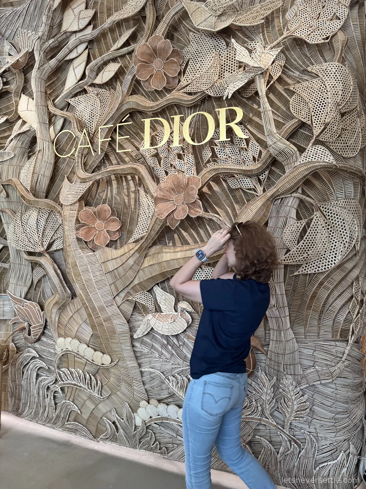 Inga reaching up to touch a floor-to-ceiling woven rattan wall of leaves, flowers and birds with the gold words Cafe Dior mounted on it