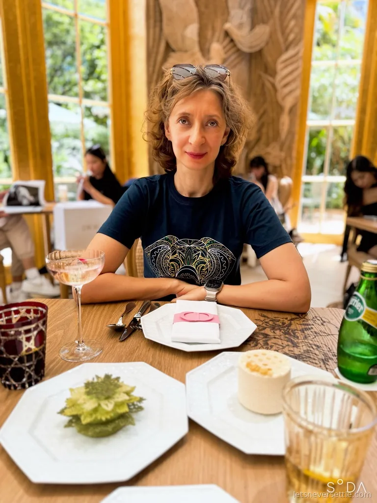 Inga at a table set with a green matcha pastry, a coupe cocktail and a bottle of Perrier, gold-framed windows and a carved wall behind