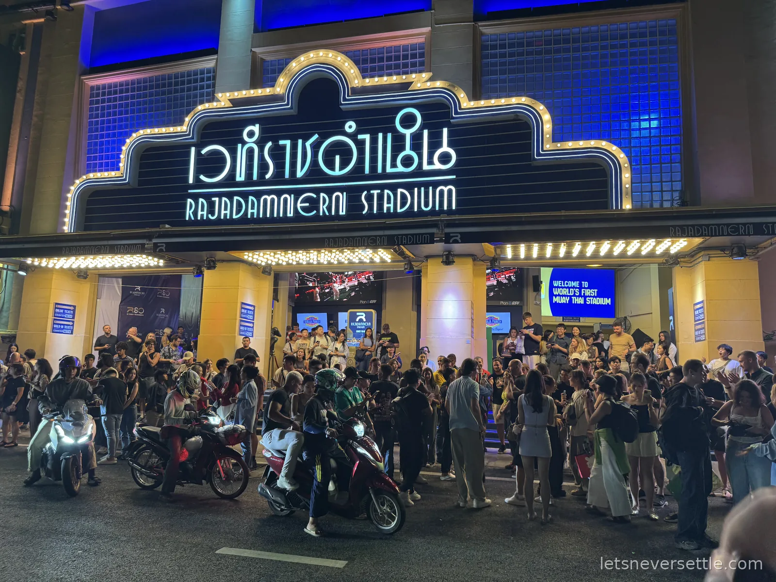 The illuminated Rajadamnern Stadium marquee at night with Thai and English lettering, crowds and parked motorbikes spilling into the street below