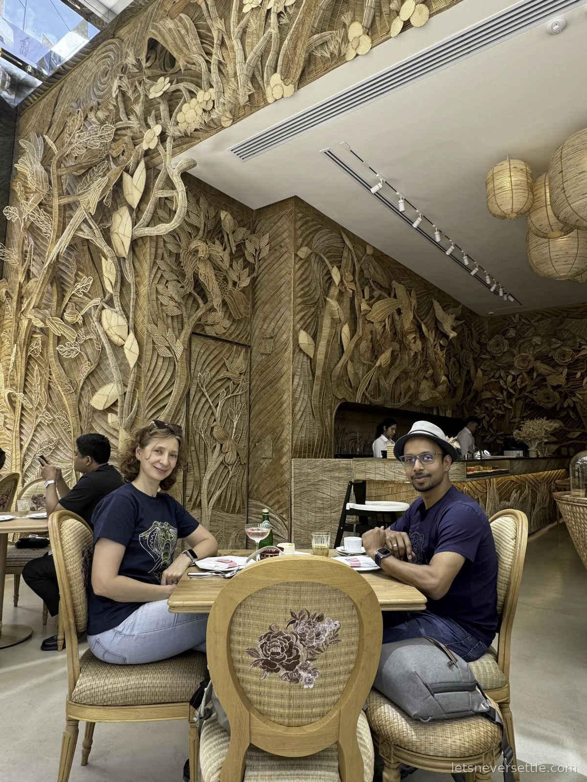 Inga and Dileep at a table in a dining room whose walls are covered floor to ceiling in hand-woven rattan reliefs of plants and birds