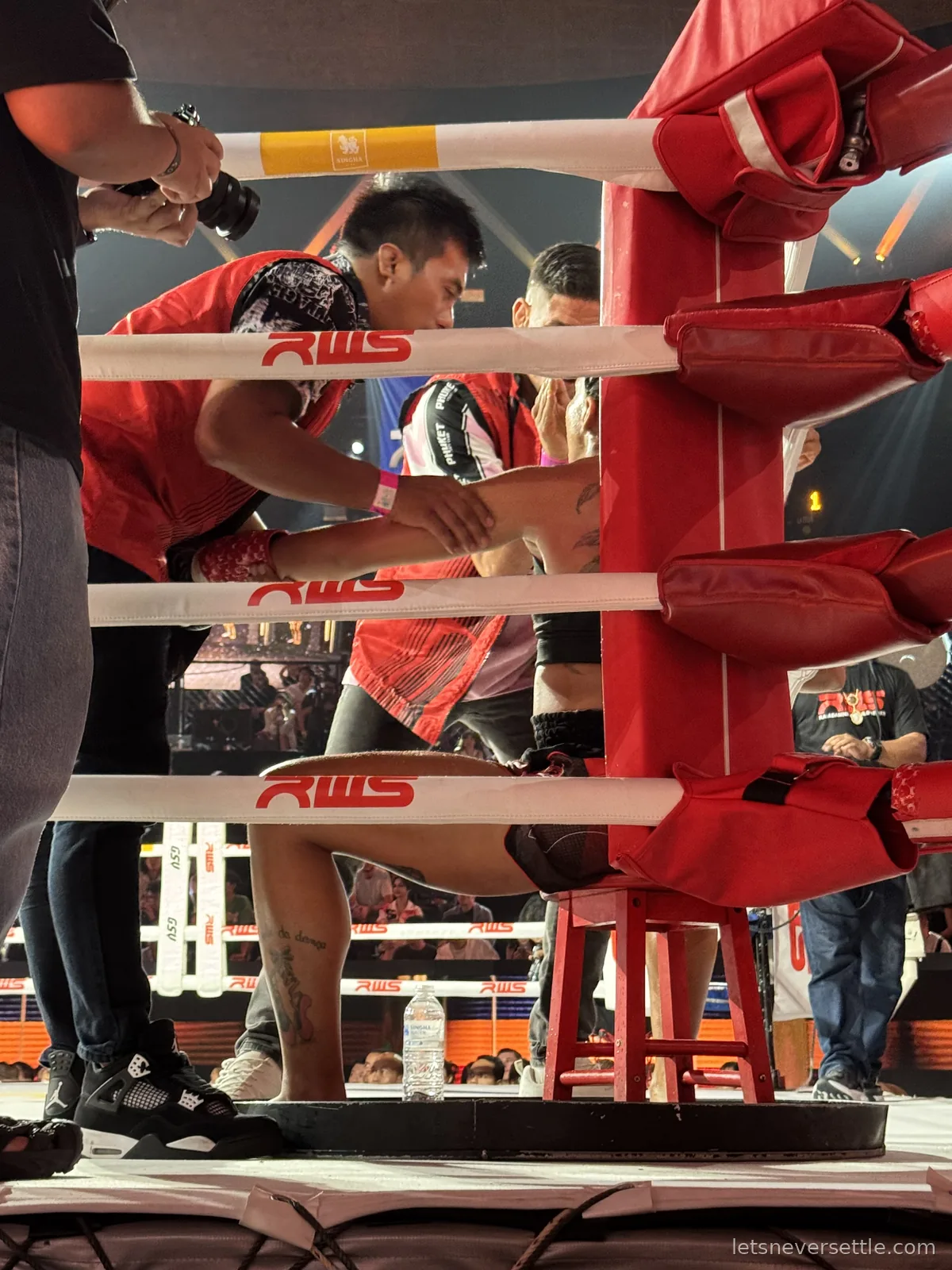 Cornermen in red vests working on a seated fighter's leg through the ropes at the red corner post between rounds