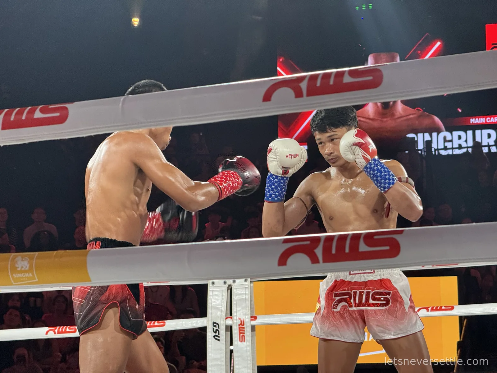 Two Muay Thai fighters squaring up in the ring, one in red and white RWS shorts with blue star-patterned gloves, seen through the ropes