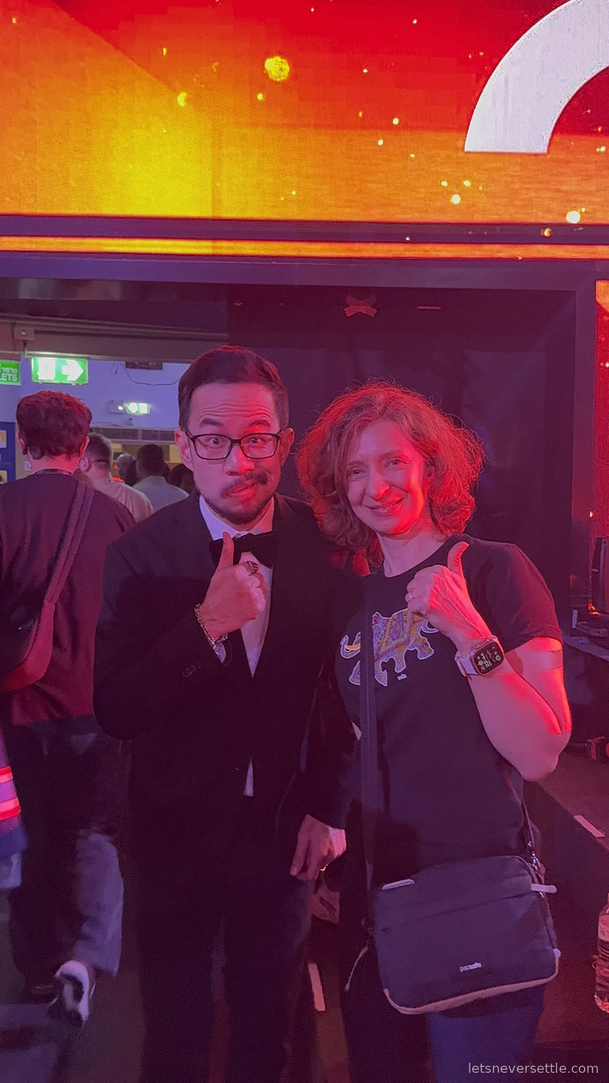 Inga giving a thumbs up next to a man in a black tuxedo and bow tie in the red-lit foyer of Rajadamnern Stadium
