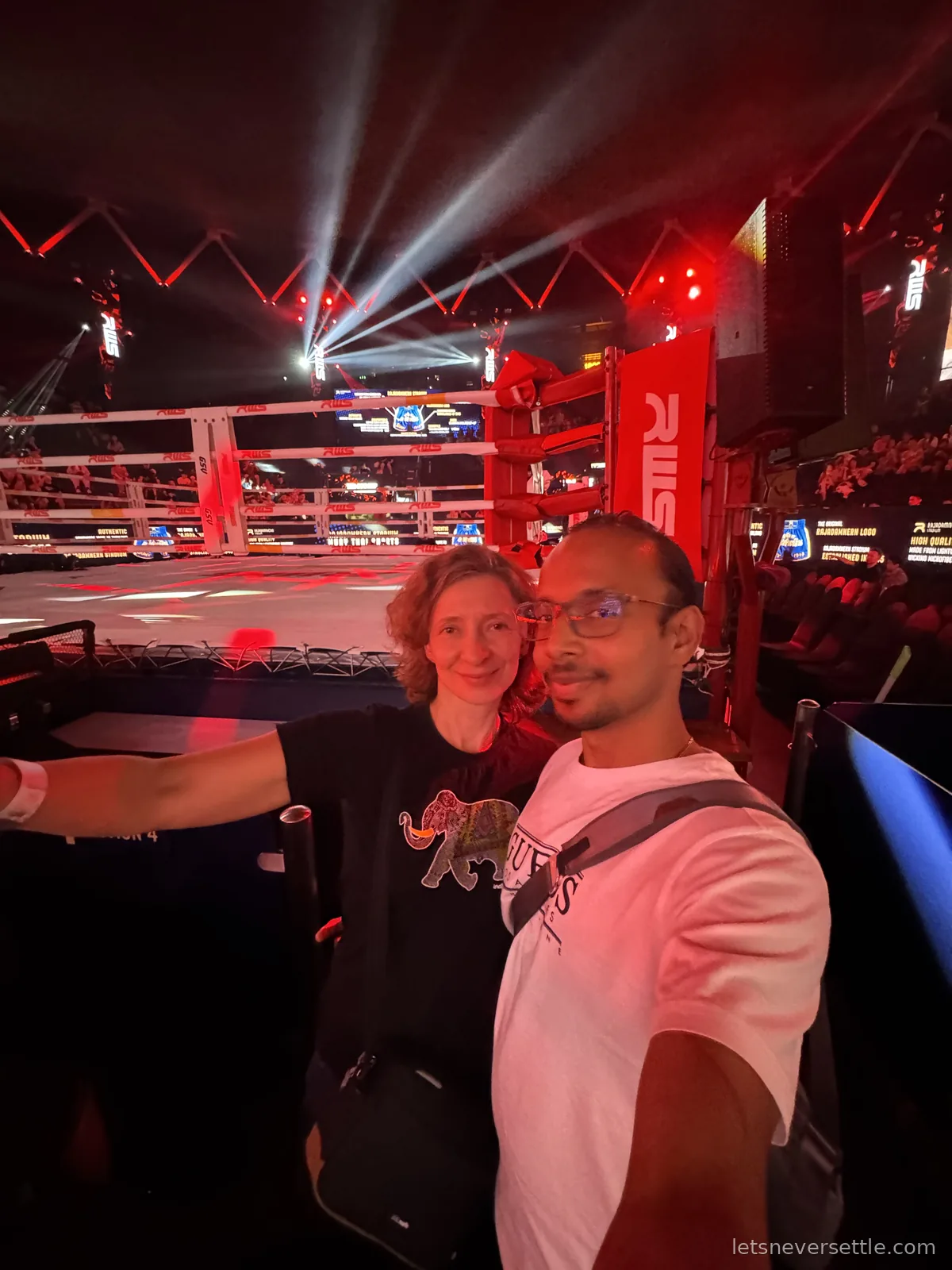 Selfie of Inga and Dileep beside the empty Rajadamnern ring before the fights, white searchlights crossing above the red RWS corner posts