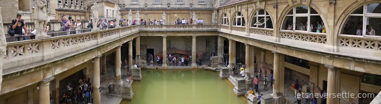 Panoramic view of the Great Bath at the Roman Baths, green water ringed by stone columns and busy terraces