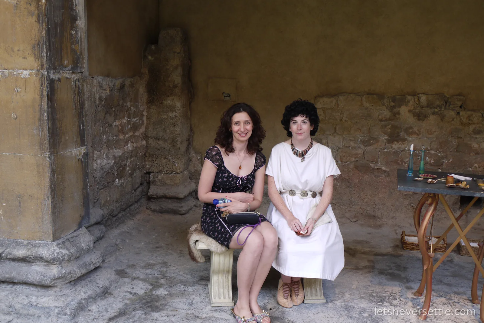 Inga sitting on a stone bench in a shaded corner of the Roman Baths beside a woman dressed in white Roman costume