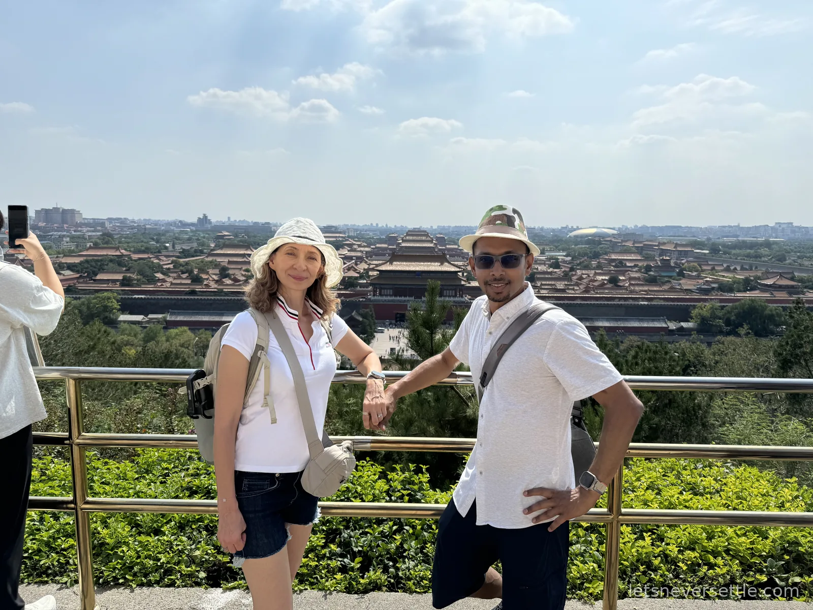 Inga and Dileep in sun hats at the Jingshan Park viewpoint railing, the Forbidden City rooftops spread out behind them