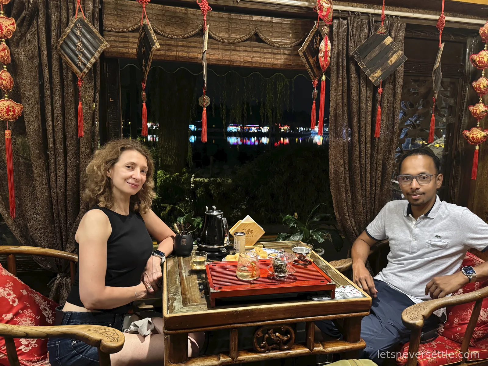 Inga and Dileep at a carved wooden table in a lantern-hung teahouse, lake lights glowing through the window behind