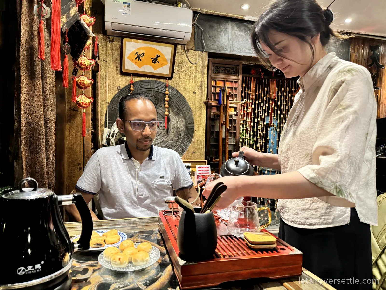 Dileep watching a hostess in traditional dress pour tea at a wooden tea tray, pastries on the table