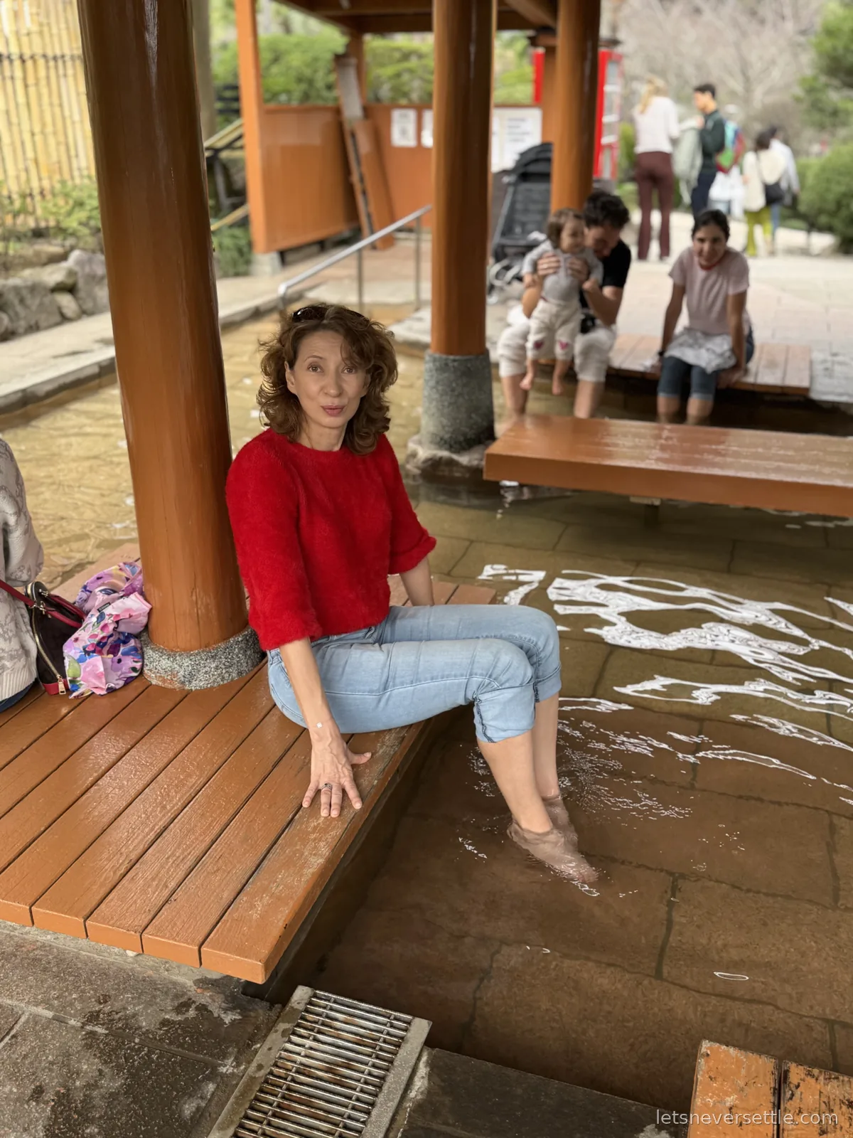 Inga in a red jumper sitting on a wooden platform with her feet in a steaming public foot bath