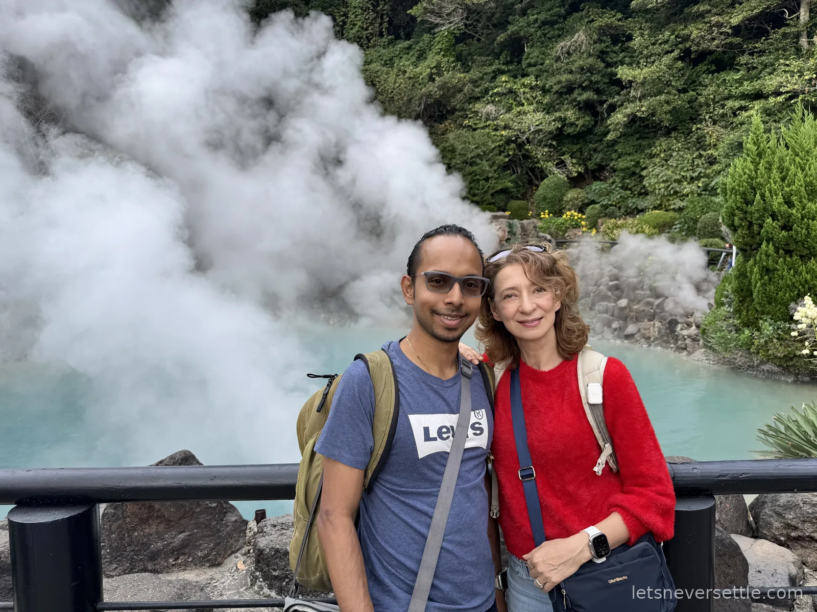 Inga and Dileep standing at a railing in front of a pale blue volcanic pond almost hidden by thick white steam