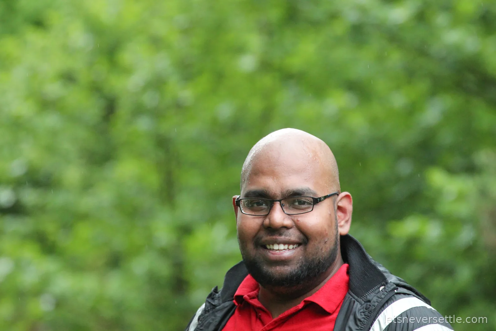 Dileep's brother in glasses, a red polo shirt and a rain jacket, smiling against a blur of wet green woodland