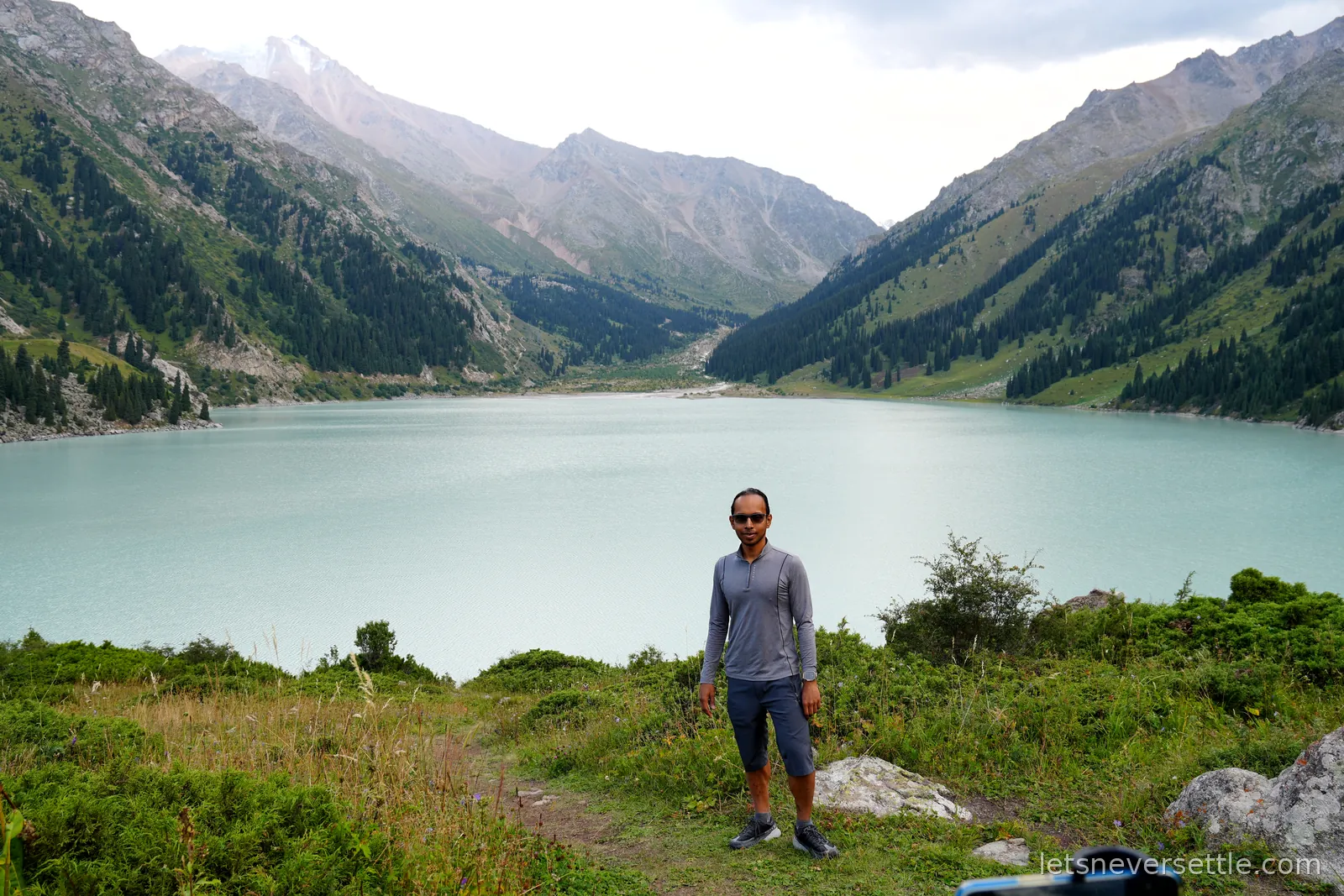 Dileep standing on the grassy bank in front of Big Almaty Lake, pale turquoise water filling the valley with bare peaks at the far end