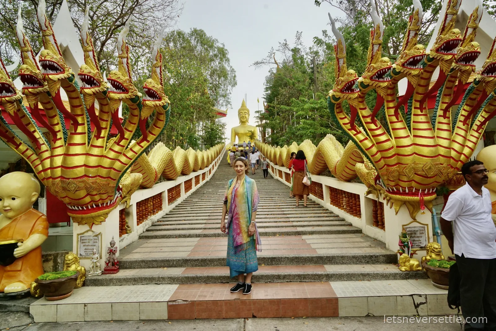 Inga in a bright printed shawl standing at the foot of the naga staircase at Big Buddha Hill, two multi-headed gold serpents rearing on either side and the golden Buddha at the top of the steps