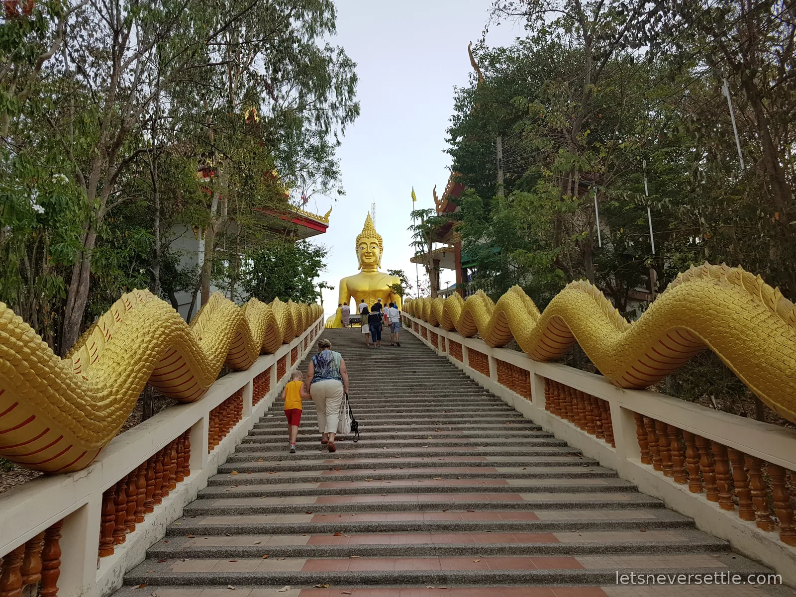 The stone staircase at Big Buddha Hill lined by the long gold scaled bodies of two naga serpents, visitors climbing towards the seated golden Buddha at the top