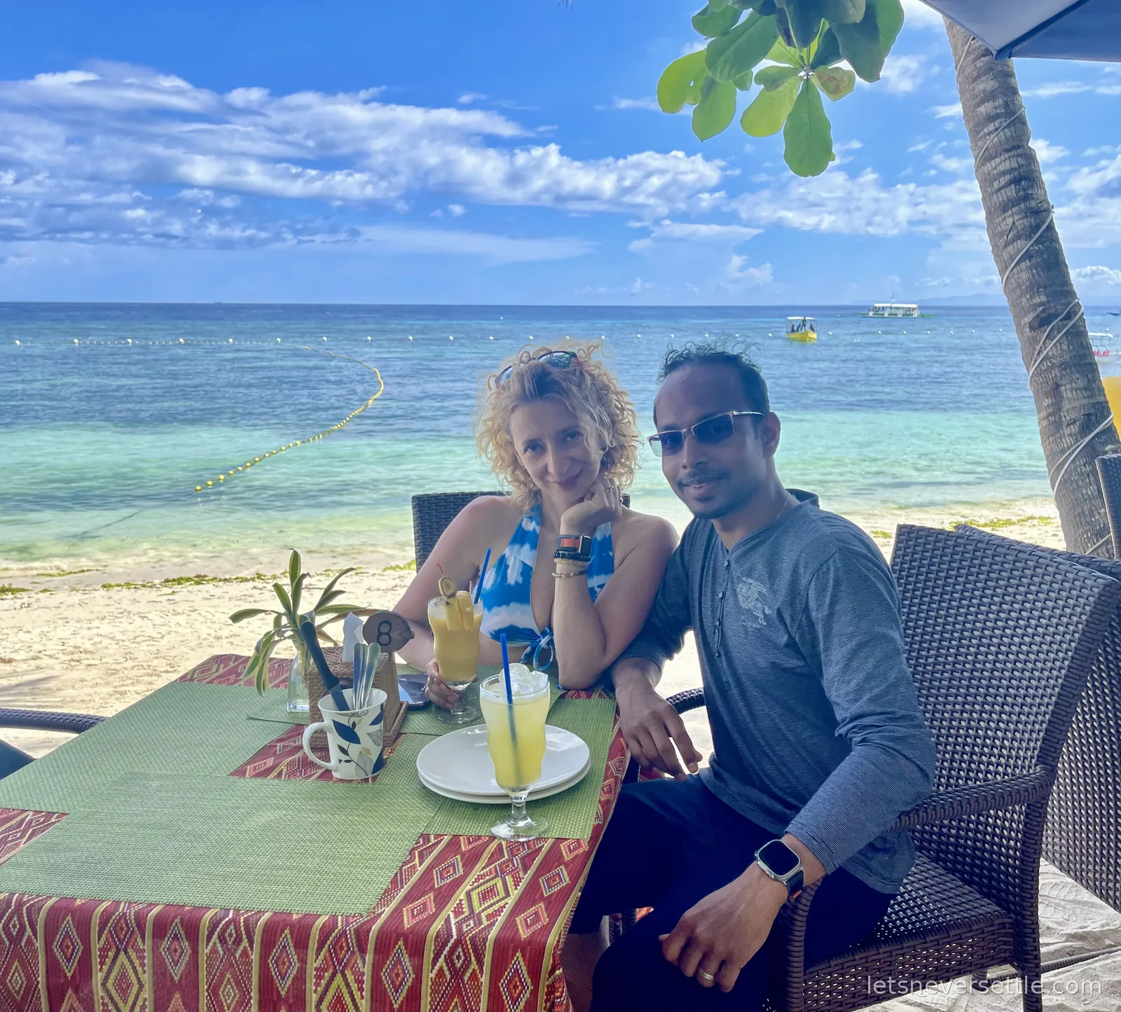 Inga and Dileep with fresh mango shakes at a beachfront table on Alona Beach, turquoise shallows and boats behind