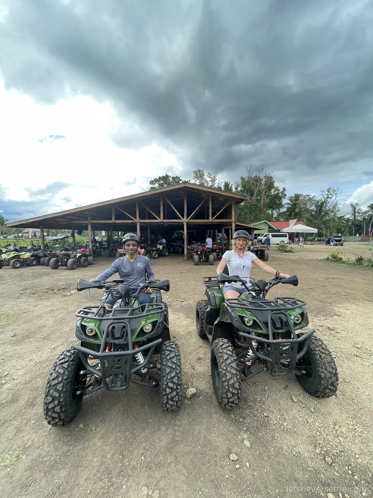 Inga and Dileep sitting on green quad bikes on a dirt track near the Chocolate Hills, storm clouds massing overhead