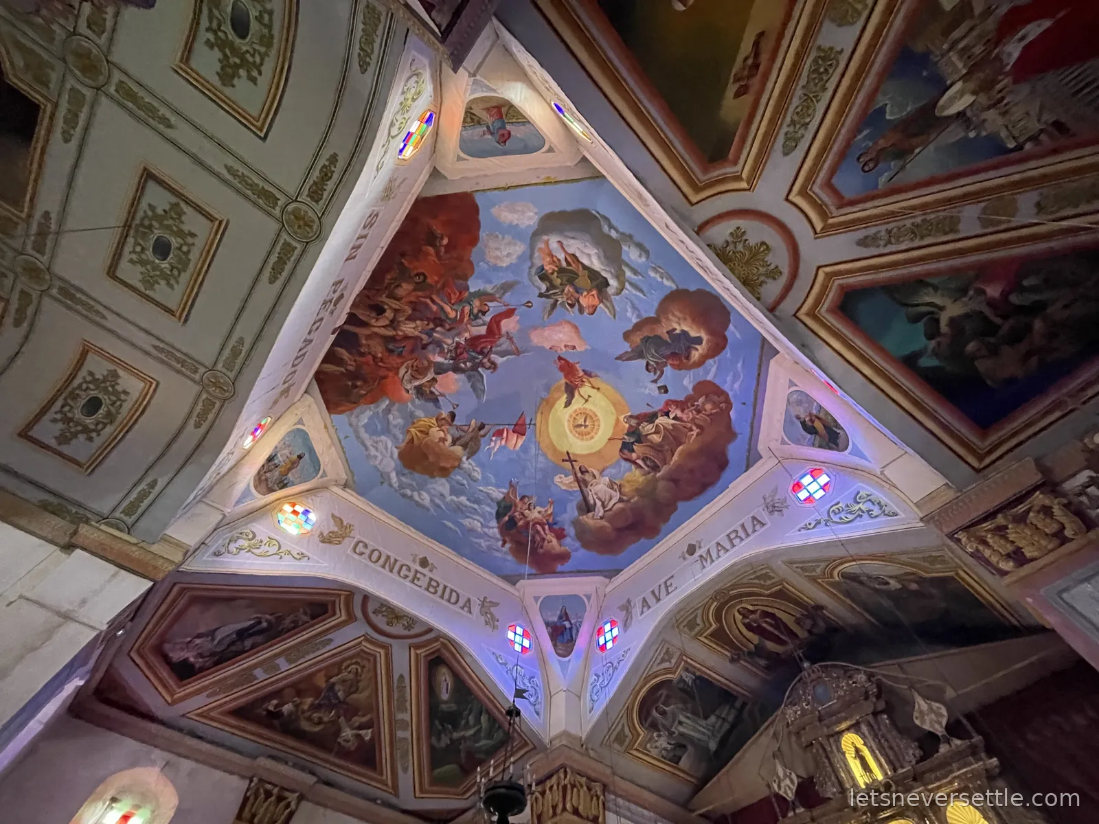The painted vaulted ceiling of Baclayon church showing biblical scenes in panels, with the Latin inscription to Mary running along the edge