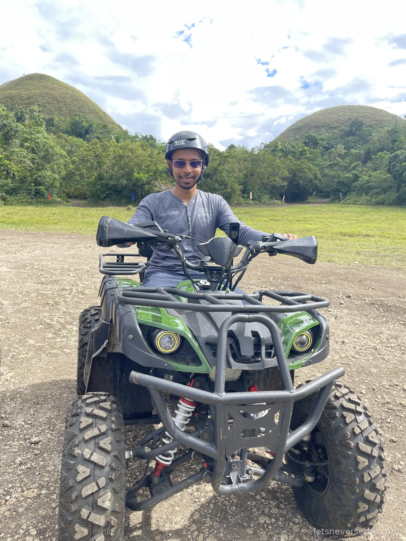 Dileep in a helmet sitting on a green and black quad bike on a dirt track with the rounded Chocolate Hills rising behind him