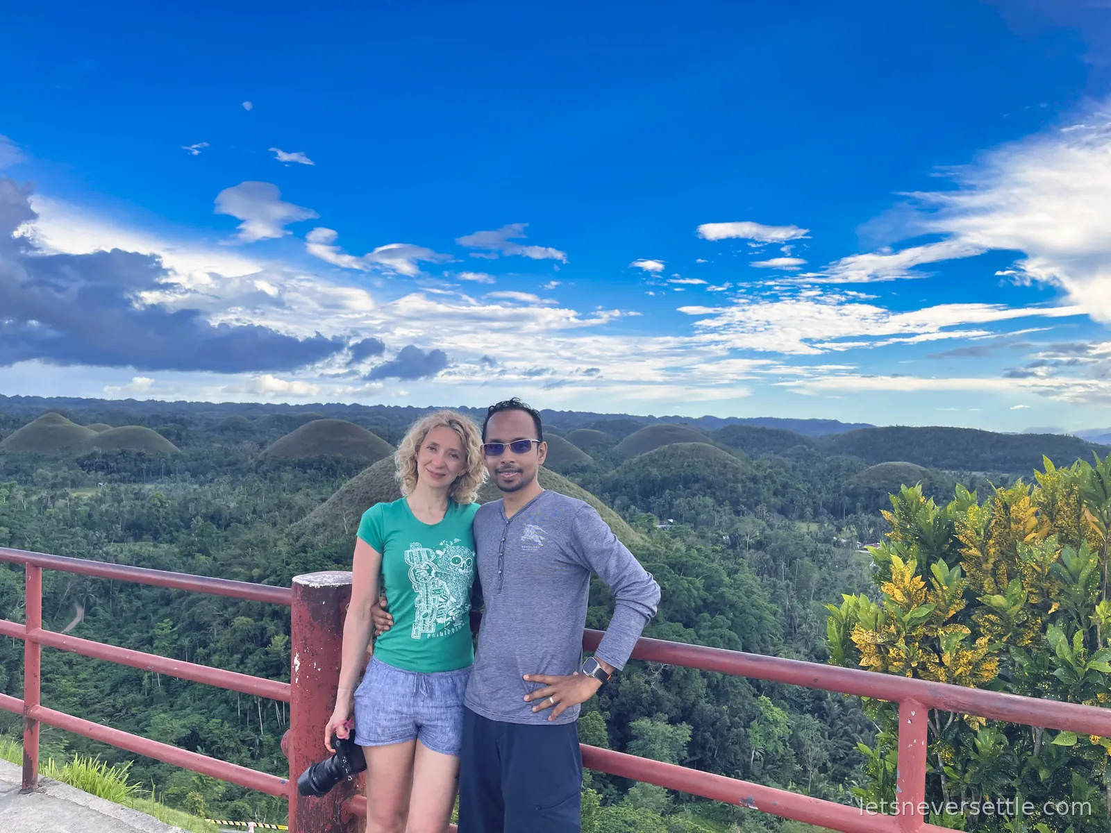 Inga and Dileep at a red railing on a viewpoint above the Chocolate Hills, green rounded hills and forest stretching to the horizon under a big sky