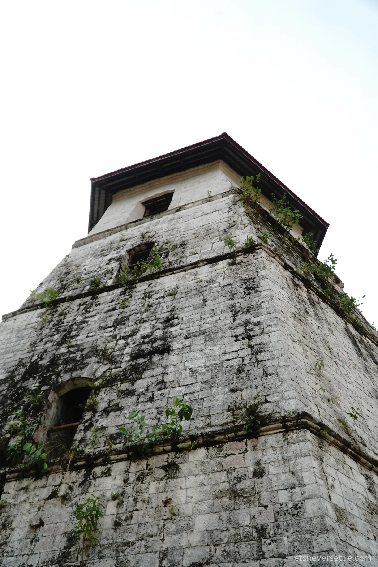A squat moss-covered coral stone bell tower on Bohol with a dark pyramid roof and small openings near the top