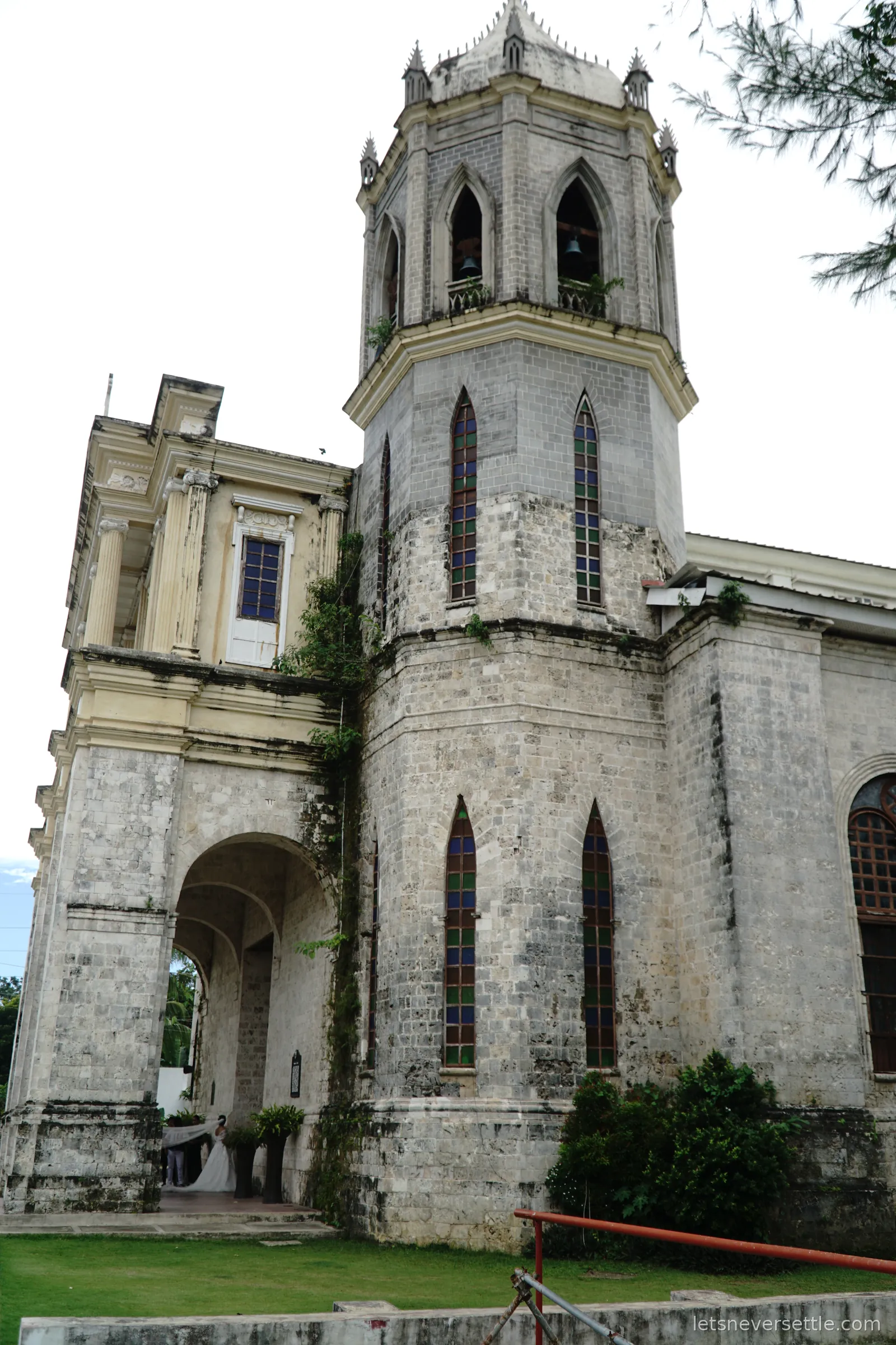A weathered grey coral stone church on Bohol with a tall bell tower and pointed spire, arched windows and moss growing on the stonework
