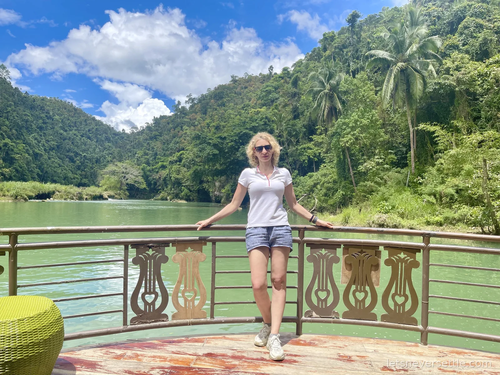 Inga leaning on the carved wooden railing of a floating restaurant deck on the green Loboc River, jungle and palms rising behind