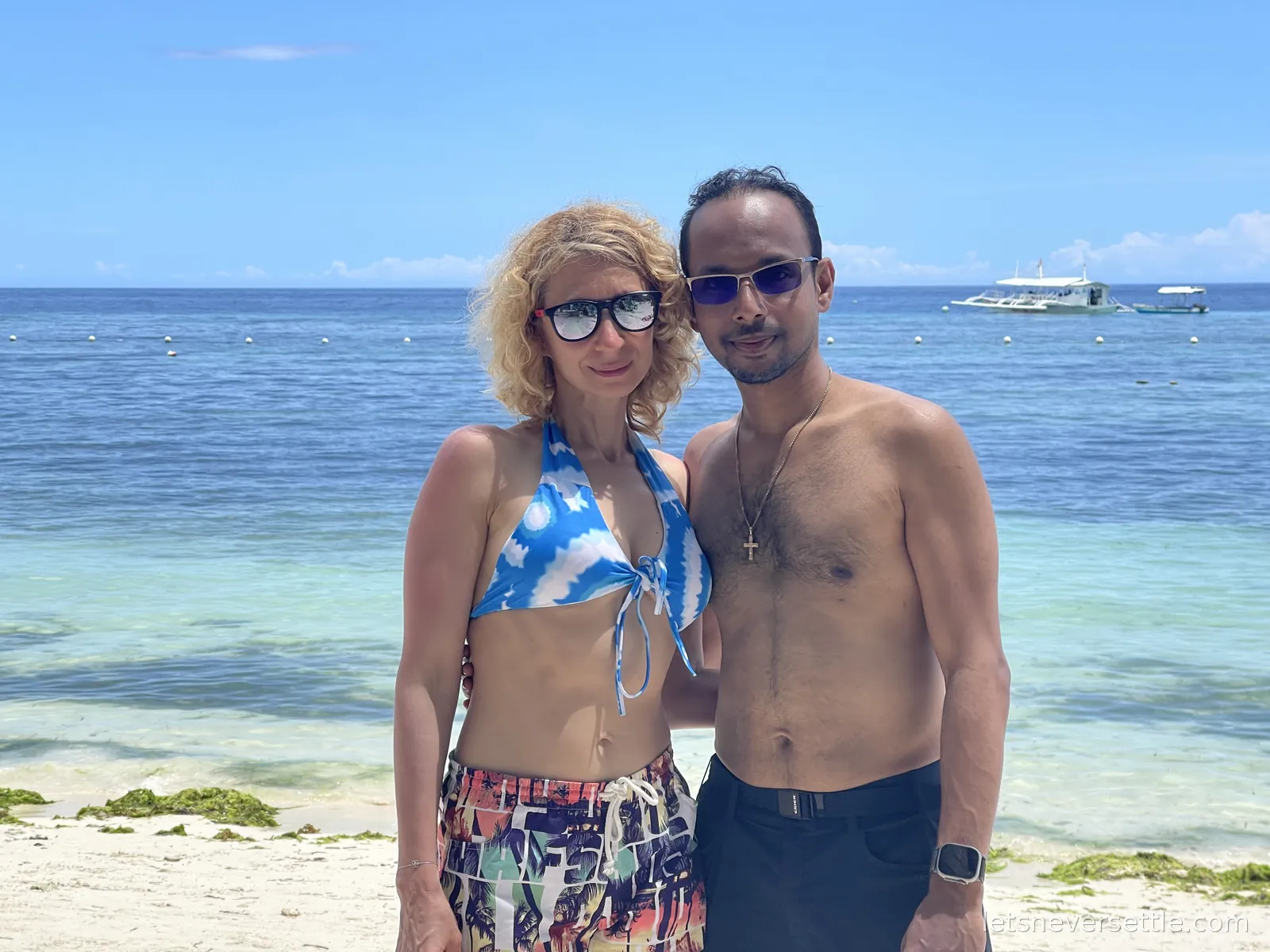 Inga and Dileep standing on a white sand beach on Panglao with clear turquoise shallows and a boat on the horizon behind them