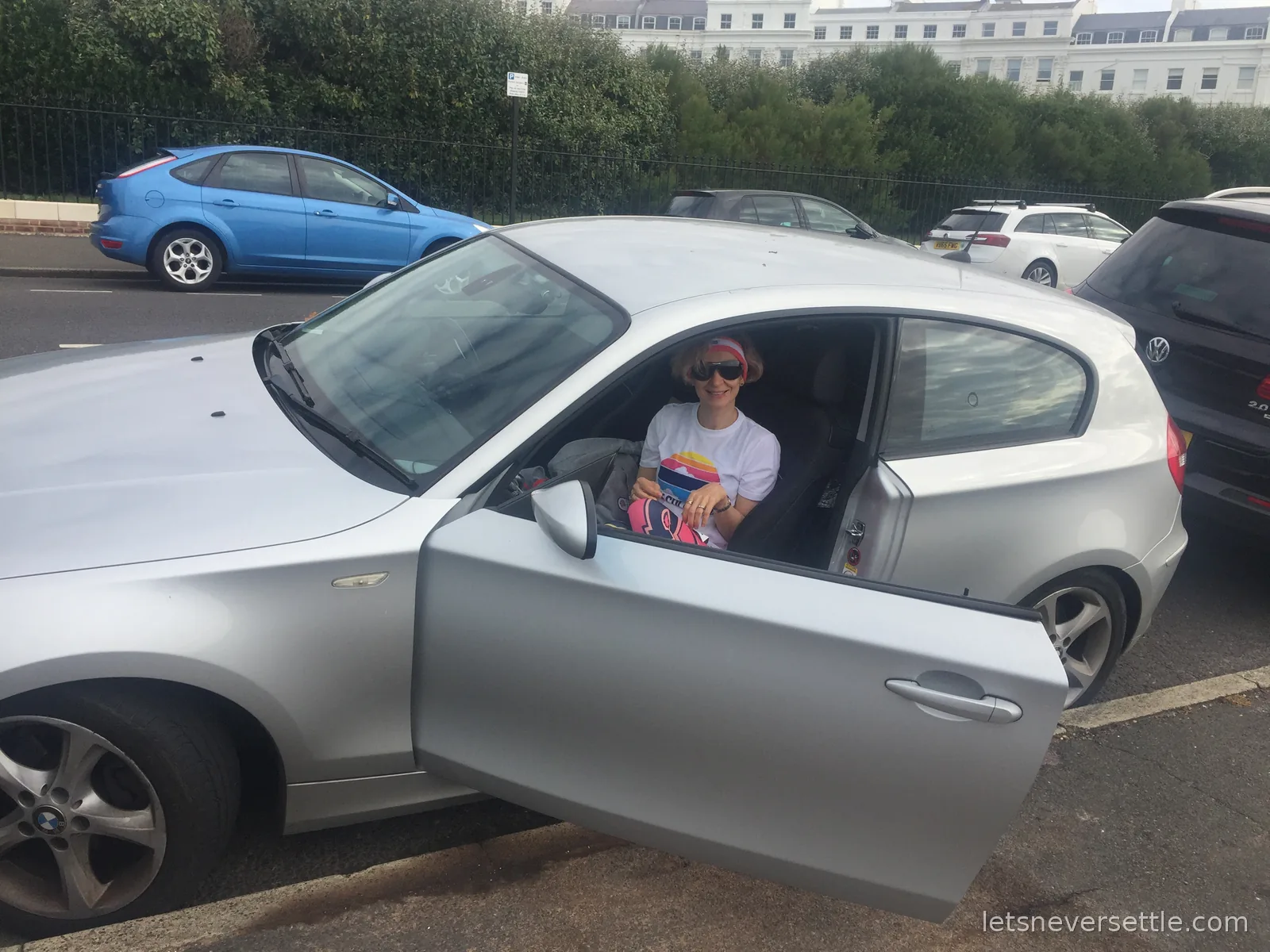 Inga in sunglasses, headband and a white event T-shirt sitting in a silver car with the door open, white seafront terraces behind