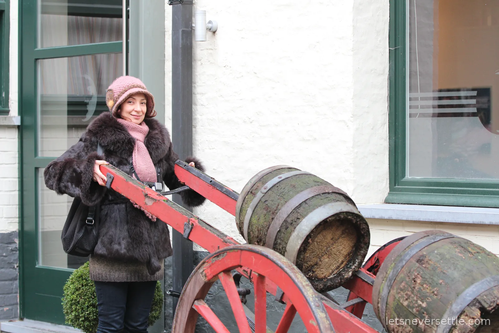 Inga in a dark fur coat and pink knitted hat standing beside an old red wooden dray cart loaded with weathered beer kegs