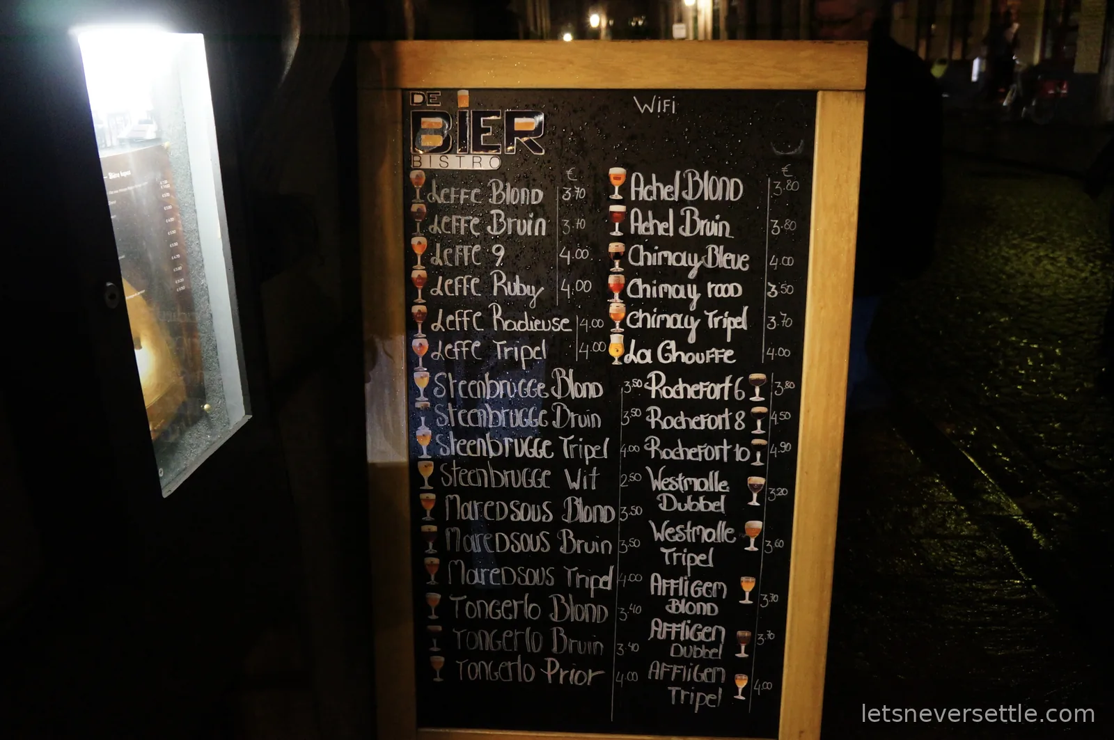 Chalkboard beer menu outside a Bruges bar at night listing Leffe, Chimay, Rochefort and Westmalle with prices