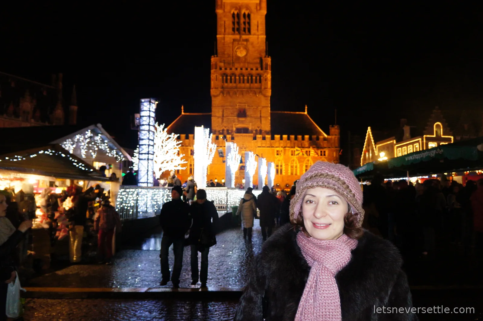 Inga in a pink knitted hat and scarf at the Bruges Christmas market at night, the illuminated Belfort tower rising behind the stalls