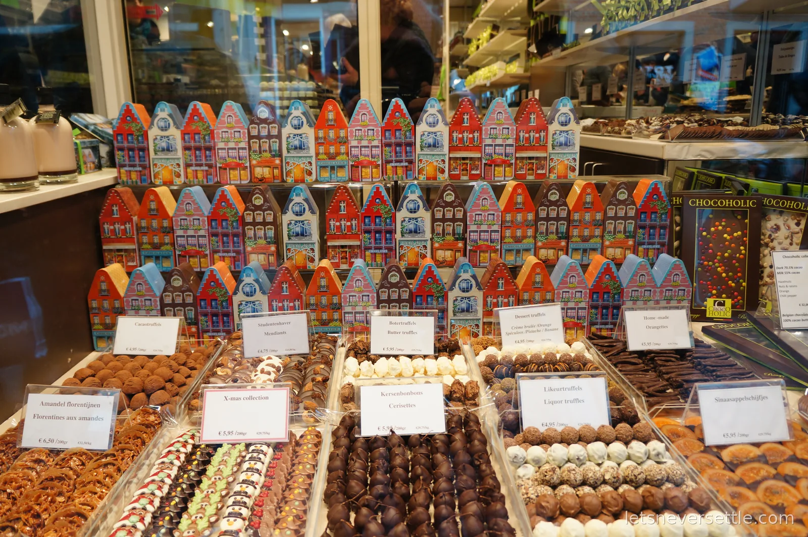 Chocolate shop display in Bruges with trays of truffles, handwritten price cards, and rows of tins shaped like Flemish gabled houses