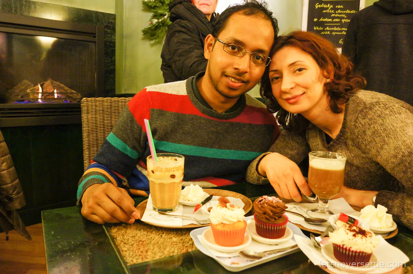 A couple leaning together at a cafe table set with three cupcakes, a latte macchiato and a coffee topped with cream