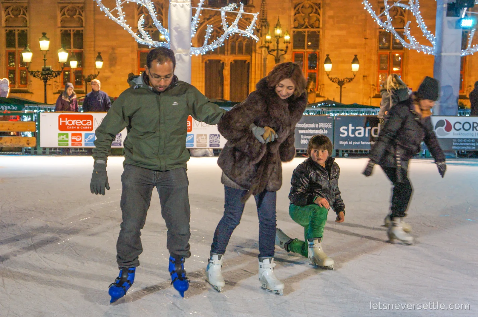 A man and a woman holding on to each other for balance on an open-air ice rink at night, with lit trees and old facades behind