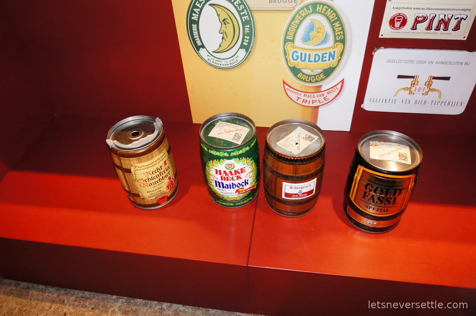 Four vintage miniature beer kegs displayed on a red shelf in front of old Bruges brewery signs and labels