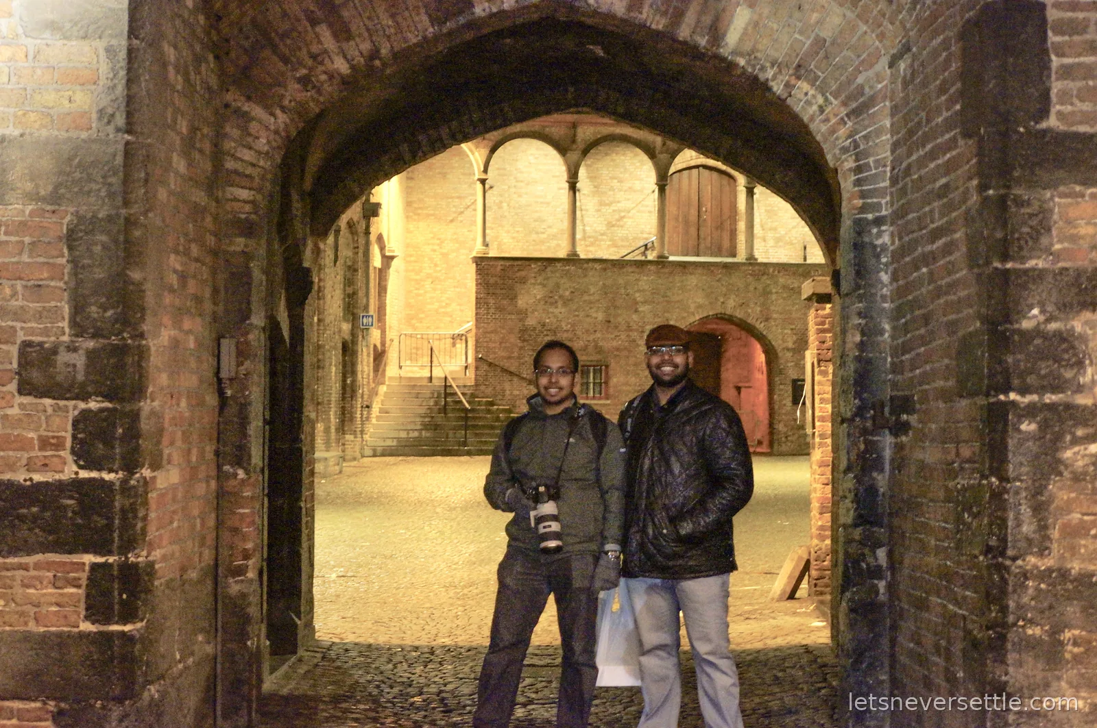 Two men standing under an illuminated brick archway in Bruges at night, one holding a DSLR camera and one in a black leather jacket