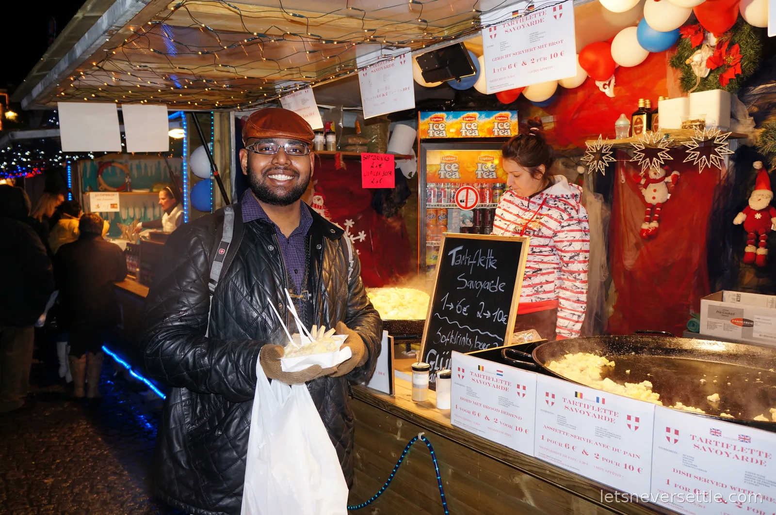 Dileep's brother in a black leather jacket and flat cap smiling with a tray of tartiflette at a Christmas market food stall