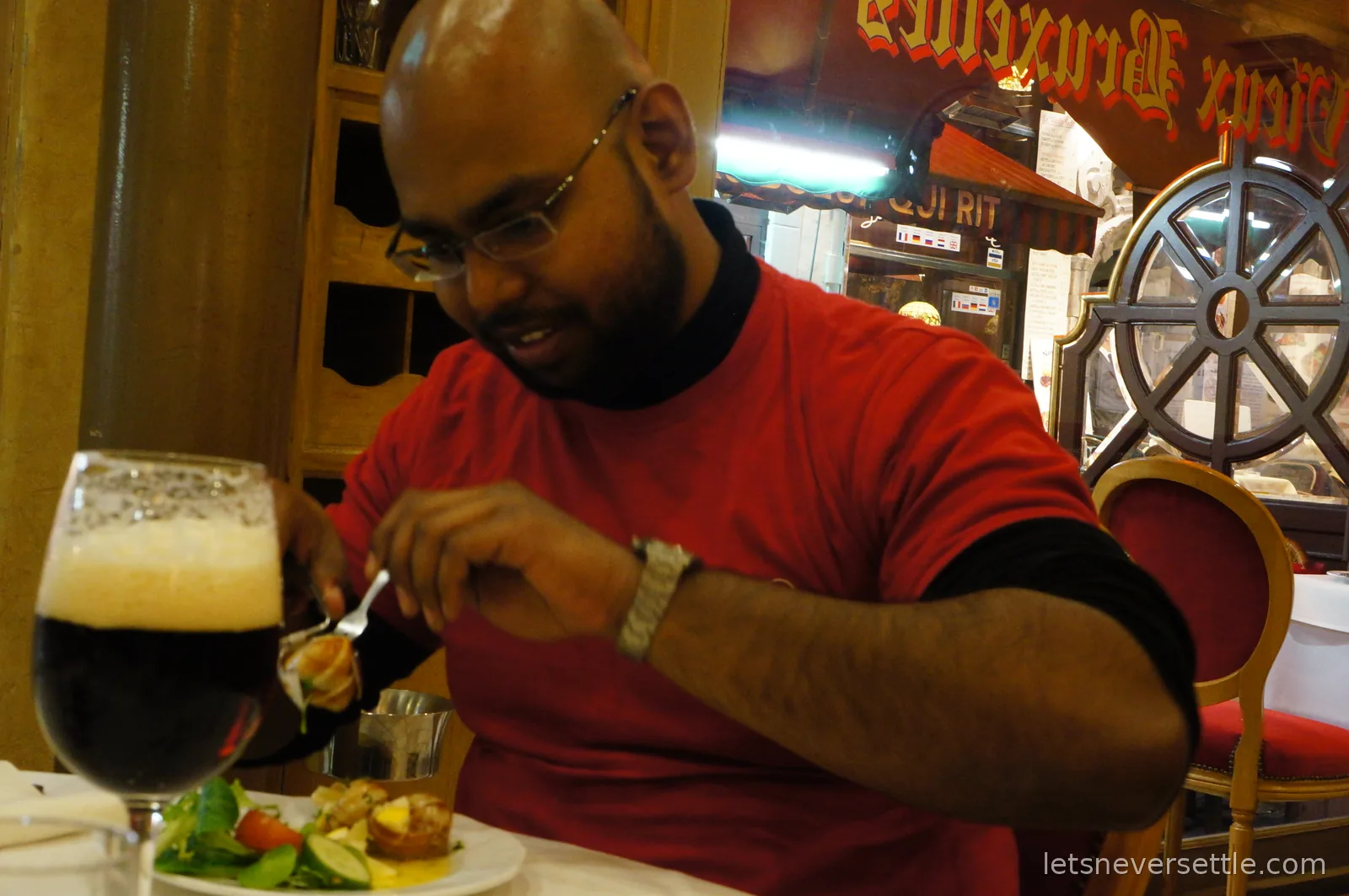 A diner in a red shirt lifting food on a fork above a plate with salad, a glass of dark beer with a thick foam head in the foreground
