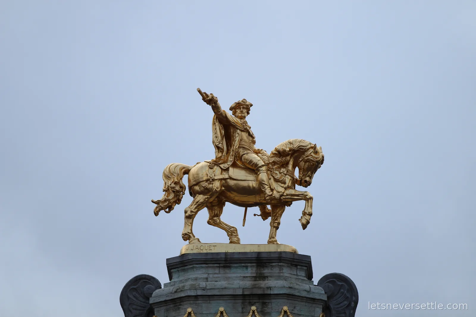 Gilded equestrian statue of a rider pointing forward on a prancing horse, mounted on a rooftop pedestal against a pale grey sky