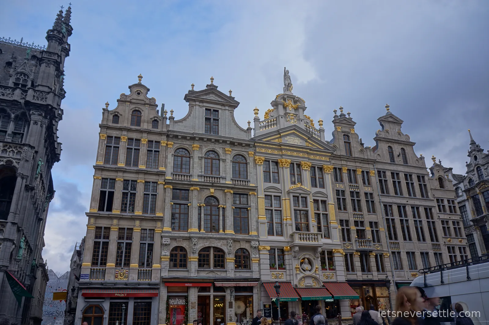 Row of ornate gilded guildhall facades on the Grand Place in Brussels under a cloudy sky, with the gothic town hall tower at the left edge