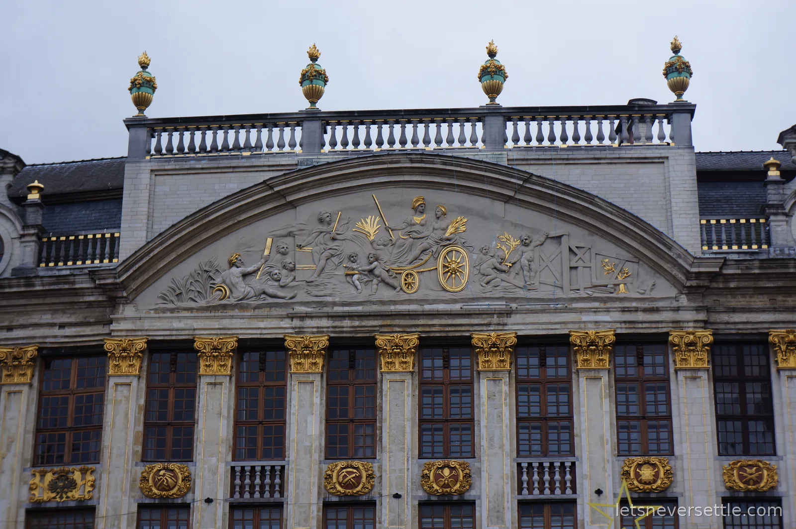 Curved guildhall pediment with a gilded relief of figures and a chariot wheel, topped by a stone balustrade and golden flame urns