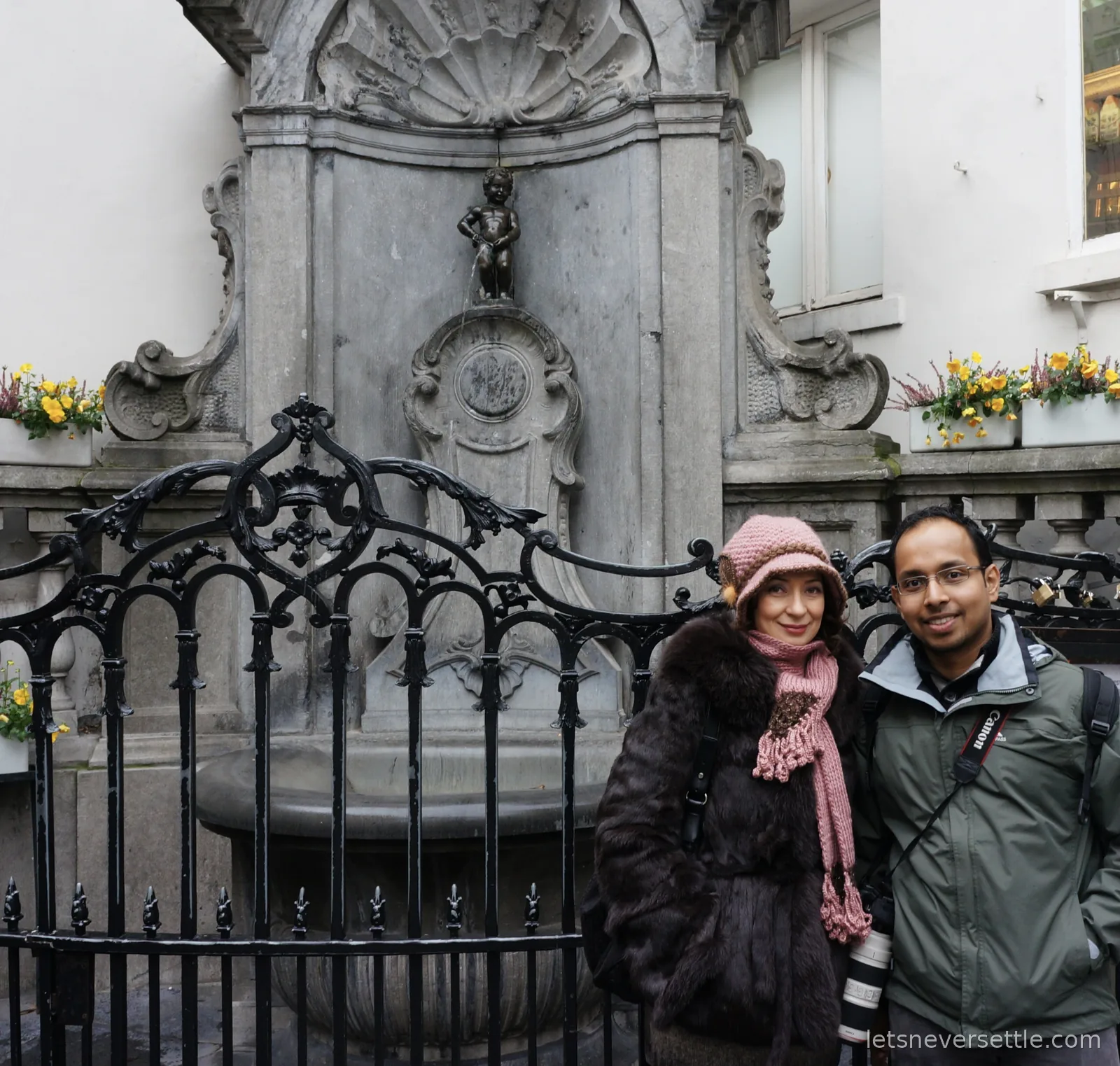 Inga in a pink knitted hat and Dileep with a camera standing at the black iron railing in front of the Manneken Pis fountain