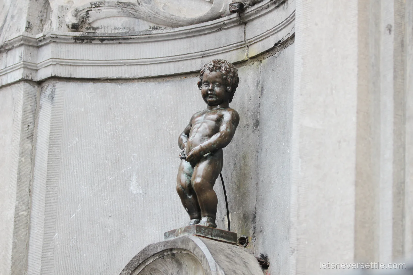 Close-up of the small bronze Manneken Pis statue mid-stream in its curved stone niche
