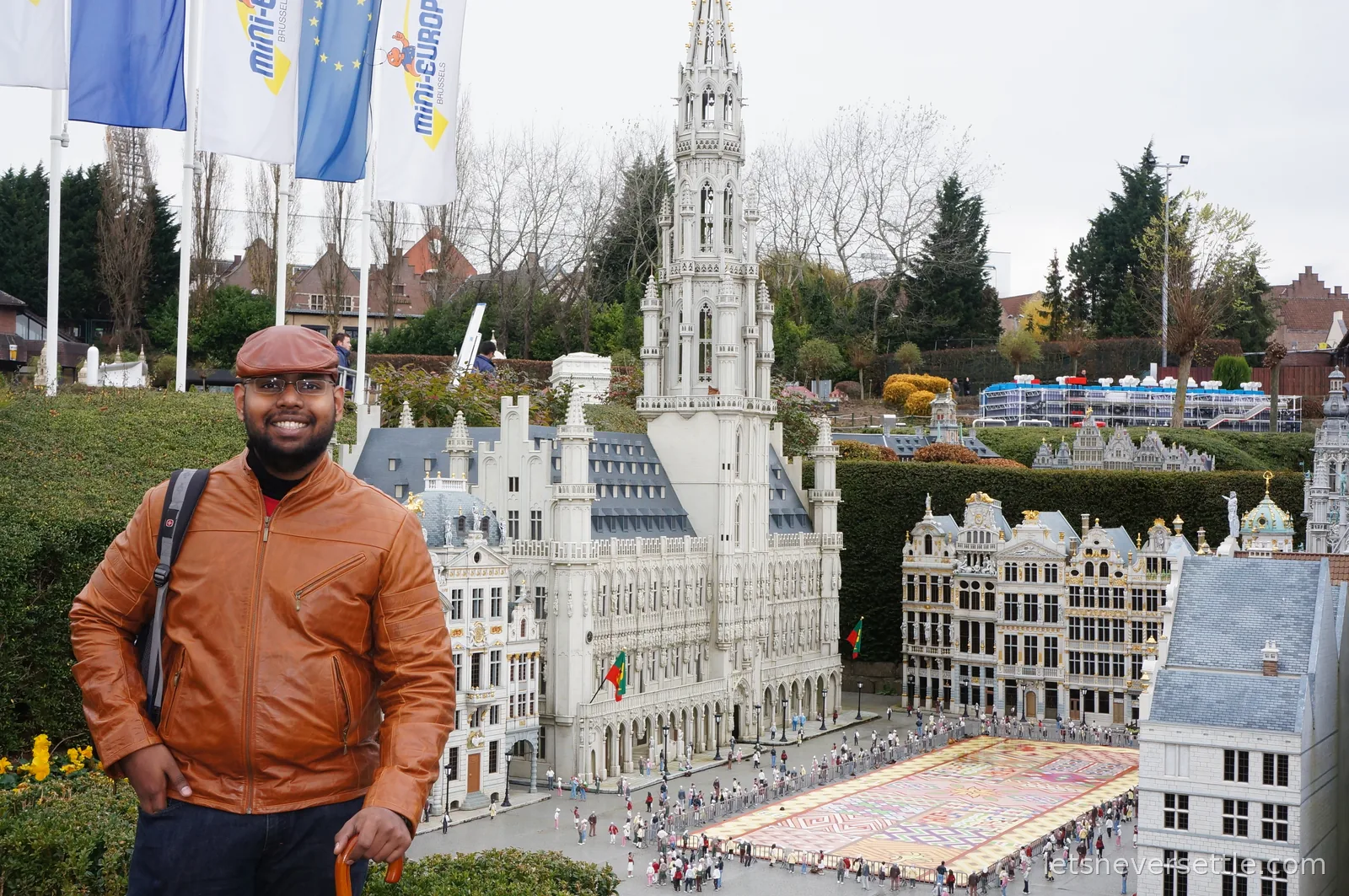 Dileep's brother in an orange leather jacket and flat cap standing beside the miniature Brussels town hall model at Mini-Europe